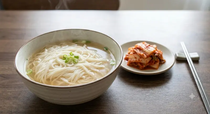 맑은 국물의 국수 한 그릇과 곁들여진 배추김치 보울, 그리고 젓가락이 놓인 심플한 상차림.
