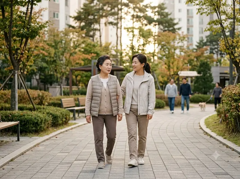 자연이 어우러진 도심 속 아파트 산책로에서 가벼운 겉옷을 입고 건강을 챙기기 위해 여유롭게 걸으며 자연스럽게 대화하고 있는 평화롭고 희망찬 모습의 모녀(또는 중년 여성들)의 사진.